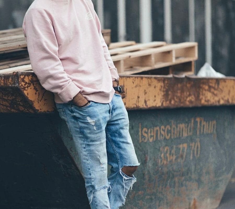 A man in a pink sweatshirt and faded blue jeans leaning against a wall
